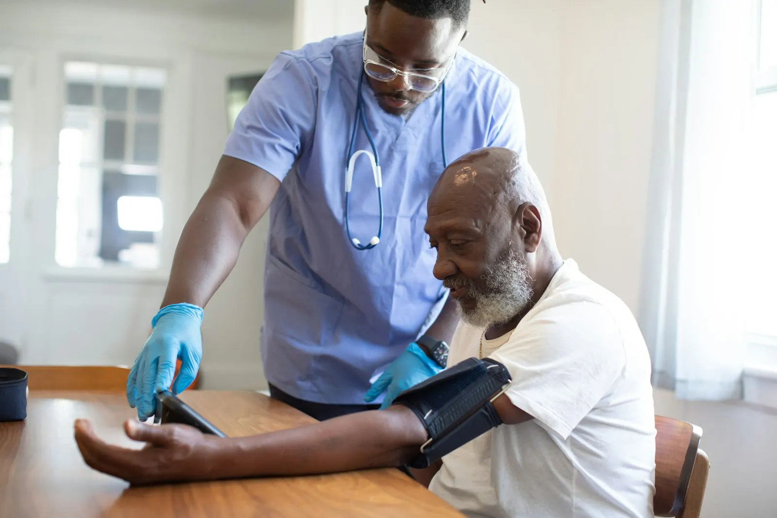A caregiver in blue scrubs checking an older man's blood pressure at home